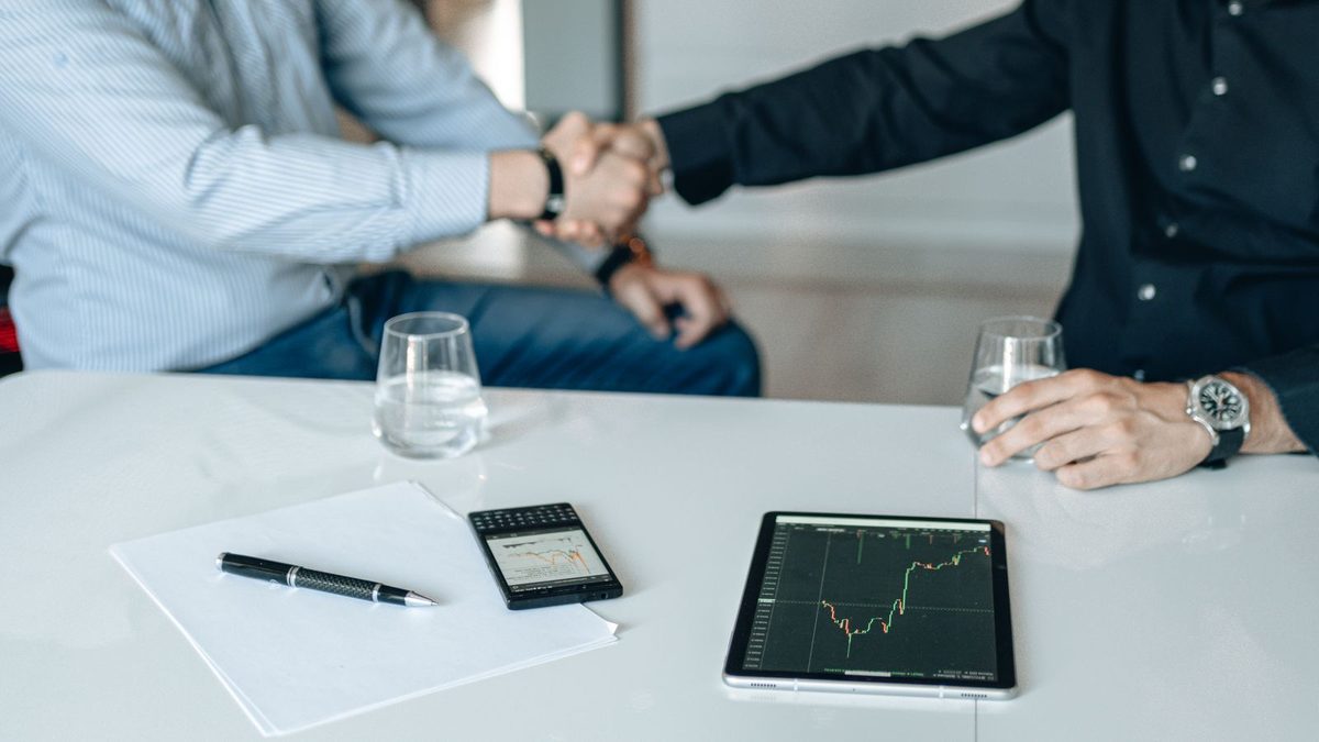 Two businesspeople shaking hands across a table with a tablet and notes